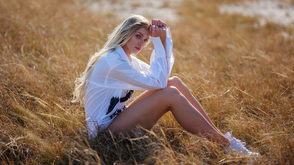 A Girl In A White Shirt Sitting In A Field Gently Locking Eyes With The Viewer Wallpaper